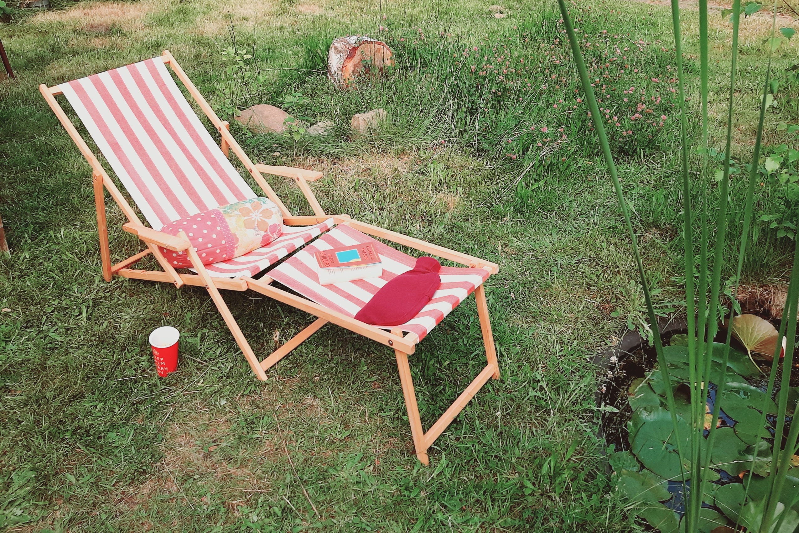 Mit rot-weiß-gestreiftem Stoff bezogener Holzliegestuhl steht auf dem Rasen an einem Gartenteich, darauf liegen eine bunte Nackenrolle, ein Buch und eine Wärmflasche, daneben steht ein roter Becher im Gras